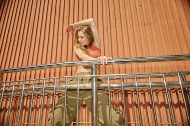 Genç bir kadın turuncu metal bir duvara karşı egzersiz yapıyor. Kolları yukarıda ve sırtı parmaklıklara yaslanmış şekilde güç ve esneklik yayıyor.