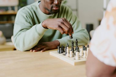 Genç siyah adam ahşap masada oturuyor ve tanınmayan arkadaşıyla satranç oynuyor.