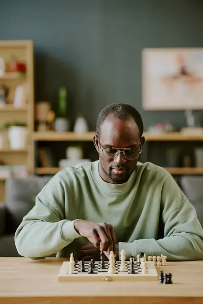 stock image Vertical shot of young African American man sitting at wooden desk in living room solving chess problem, copy space