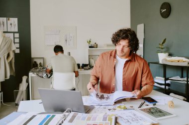 Moda tasarımcısı, arka planda dikiş makinesi kullanırken albüm sayfalarını kıyafet taslaklarıyla çeviriyor.
