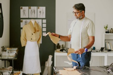 Olgun Hintli terzi elinde iki kumaş kalıbıyla modern atölyede onlara bakıyor.