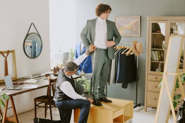 Bir dizinin üstünde duran ve aynaya bakarken kıvırcık saçlı modelin ölçümlerini yapan terzi.