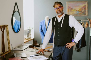 Mature Indian tailor dressed in uniform leaning on antique wooden table while posing for portrait clipart