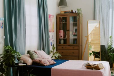 Ev bitkilerinin olduğu rahat bir yatak odası ve kitaplarla dolu bir kitaplık ve dekoratif malzemelerle dolu, mavi perdeli pencerelerden akan güneş ışığıyla yıkanmış bir kitaplık.