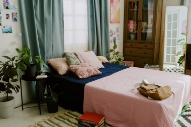 Pastel temalı dekorlu, bitkilerin, kitapların ve yumuşak tekstil ürünlerinin vurgulandığı büyüleyici bir yatak odası. Yatak yastık ve battaniye ile süslenir, davetkar atmosfer yaratır.