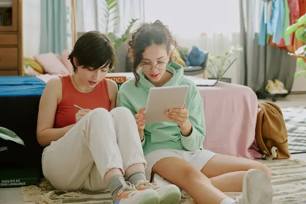 stock image Two young women collaborating on tablet while sitting on floor in cozy living room, one is writing in notebook, both focused on work