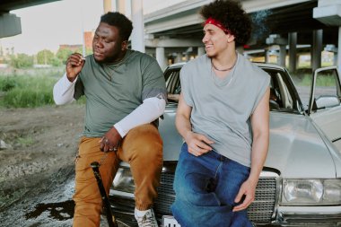 Çift ırklı arkadaşlar eski arabanın yanında vakit geçirirken sert çocuk sigara içiyor.