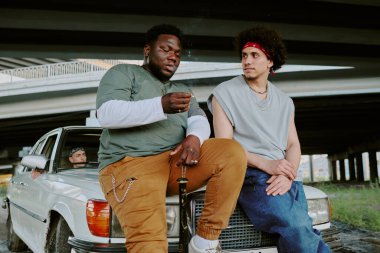 Çift ırklı eski dostlar birbirleriyle iletişim kurarken beyaz arabaya yaslanıyorlar.