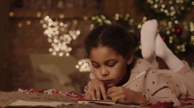Yakın plan, yerde yatan melez bir kız. Noel Baba 'ya mektup yazıyor. İçinde pırıl pırıl ışıklar ve tatil için süslemeler var.