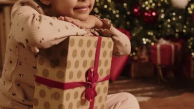 Hediye kutusuyla bacak bacak üstüne atmış, mutlu, melez bir kızın portresi. Parıldayan Noel atmosferinde kameraya poz verip gülümsüyor.