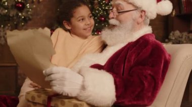 Noel Baba 'nın koltukta oturmuş, melez genç bir kızla dilek dilerken ve Noel hediyesi beklerken çekilmiş orta boy bir fotoğrafı.
