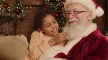 Mutlu çift ırklı bir kızın neşeli Noel Baba 'yla koltukta oturup Noel için özel dileklerle mektup okuduğu orta boy bir fotoğraf. Beyaz sakala dokunuyor.