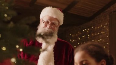 Orta boy melez kız ve Noel Baba kostümü giyen yaşlı beyaz adamın çam dallarına kırmızı parlak mücevherler koyarak Noel ağacını süslemesi.