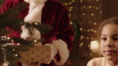 Noel Baba kostümü giymiş, beyaz eldivenli, tanınmayan bir adamın orta kesiminde melez bir kızın Noel ağacını dikkatlice süslemesine kırmızı, pırıl pırıl mücevherler veriliyor.