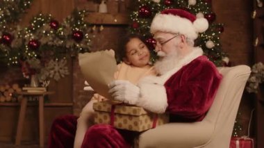 Orta yaşlı, beyaz, Noel Baba kostümü giymiş bir adam kucağında melez bir kızla koltukta oturuyor, mektup okuyor ve Noel dileklerini ve umutlarını tartışıyor.