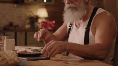 Noel şapkalı kıdemli beyaz adamın orta boy yakın çekimi mutfakta masada oturmuş akıllı telefondaki mesajları ya da e-postaları kontrol ediyor, kahve içiyor ve yemek yiyor.