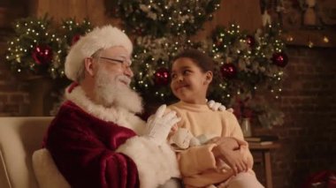 Noel Baba kostümü giymiş yaşlı bir adamın portresi ve elinde yumuşak bir oyuncak tutan melez bir kız Noel atmosferinde birlikte oturuyor, gülümsüyor ve kameraya bakıyor.