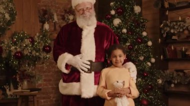Noel Baba kostümü giyen yaşlı beyaz adamın portresi ve dekore edilmiş odada kameraya poz veren melez bir genç kız. Noel ağacı ve arka planda yanıp sönen ışıklar