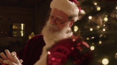 Noel Baba kostümü giyen yaşlı beyaz adamın Noel ağacını parlak kırmızı mücevherlerle süsleyerek festival sezonuna hazırlandığı orta boy yakın çekim.