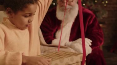 Mutlu, melez bir genç kızın Noel hediyesiyle kutusunu açtığı, Noel Baba 'ya sarılıp teşekkür ettiği, arka planda ışıldayan neşeli bir sahne.