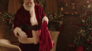 Yaşlı beyaz adamın Noel Baba kılığına girip hediye kutularını kırmızı torbaya koyduğu, arka plandaki Noel kutlamasına hazırlandığı orta boy bir fotoğraf.