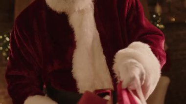 70 'li yaşlarda, Noel Baba kostümü ve gözlük takmış, hediye kutularını kırmızı torbaya koyup Noel gecesine hazırlanan beyaz bir adamın yakın çekimi.