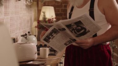 Kafkasyalı kıdemli bir adamın, Noel Baba rolüne hazırlanırken, rahat bir mutfakta durup, ekmek kızartırken, Noel gazetesi okuduğu yakın çekim görüntüleri.