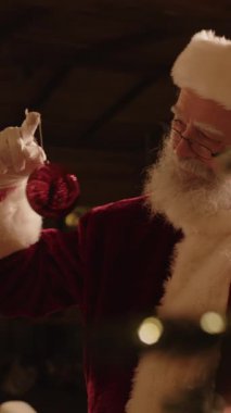 Noel Baba kostümü giyen yaşlı beyaz adamın dikey yakın çekimi. Noel ağacını kırmızı parlak mücevherlerle süslüyor. Festival sezonuna hazırlanıyor.