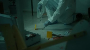 Koruyucu üniformalı profesyonel bir kriminalist olay yerinde kan lekeleri, bıçak ve kanıt işaretleri olan dağınık zeminden mermi kovanlarını plastik torbaya dolduruyor.