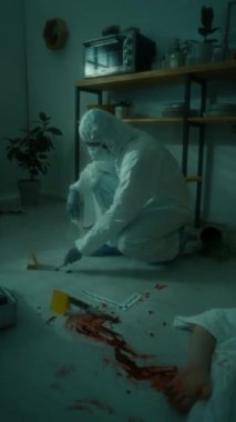 Koruyucu takım elbiseli, maskeli ve eldivenli adli tıp teknisyeninin karanlık odadaki cinayet mahallinde plastik poşette kanıt topladığı dikey bir fotoğraf.