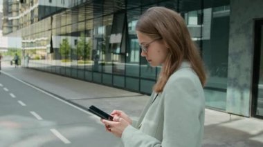 Resmi kıyafetli beyaz bir kız akıllı telefonuyla gelen mesajlara cevap verirken dışarıda panoramik pencereli büyük binanın yanında iş ortağı bekliyor.