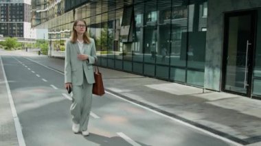 Telefon ve çanta taşıyan, takım elbiseli genç bir iş kadını ofis binasına giderken şehir caddesinde yürüyor.