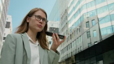 Formalite giymiş genç bir bayan hoparlör kullanırken ve dışarıda iş arkadaşlarıyla konuşurken kendi önünde akıllı telefon tutuyor.