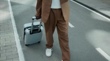 Kahverengi takım elbiseli, gözlüklü, kablosuz kulaklıklı ve tekerlekli bavullu Afro-Amerikan delikanlıyı yukarı kaldır. İş gezisinde şehir caddesinde yürürken akıllı telefon kullanarak yerini kontrol et.