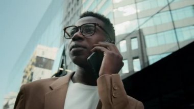Kahverengi ceketli Afro-Amerikalı genç işadamının ofis binalarının yakınındaki akıllı telefondan yaptığı konuşmayı yakından izleyin.