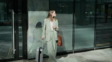 Açık renk takım elbiseli genç bir bayanın otelin yanında el çantası ve bavuluyla iş ortağı beklerken saatine baktığı uzun bir manzara.