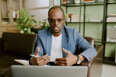Ofiste dizüstü bilgisayarda çalışan ciddi Afrikalı Amerikalı iş adamının portresi, sanal toplantıya katılırken notlar ve hareketler alıyor.