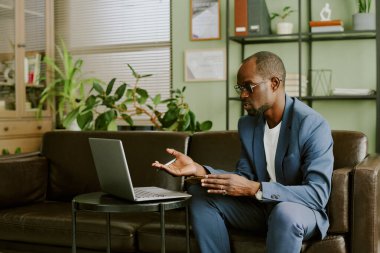 Takım elbiseli ve gözlüklü bir adam kahverengi koltukta otururken modern ofis ortamında sanal toplantı yapıyor. Çiçekler ve duvar süslemeleriyle donatılmış bir oda.