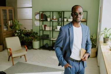 Modern ofis ortamında elinde kitaplarla duran Asyalı bir adam. Kitap rafları, koltuklar ve masa bitkileriyle iyi organize edilmiş ofis dekorasyonu.