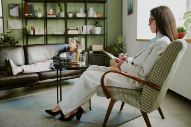 Modern ofisteki terapist, genç müşteriyle koltukta uzanmış, rahatlıyor şık bir ortamda, kitap raflarında ve arka planda görünen kapalı alandaki bitkilerin arasında.