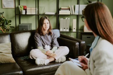 Terapi seansında danışman ile konuşurken kanepede oturan kız. Ofisin içinde arka planda kitaplıklar var.