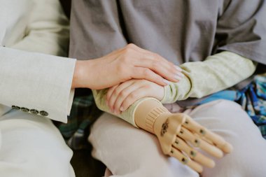 İki bireyin el ele tutuşması, rahatlık ve destek sunması. Bir kişinin protez bacağı var. Bağlantı ve özen gösterme anı.