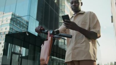 Düşük açılı, kafası karışmış, elektrikli scooterlı genç bir adamın şehir uzayındaki akıllı telefonundaki haritalarla etrafa bakınışı.