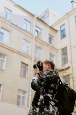 Erkek fotoğrafçı dışarıda duruyor ve şehir manzarasının fotoğrafını çekerken elinde kamera var. Arkaplan şehir çevresindeki uzun, açık renkli binalardan oluşuyor