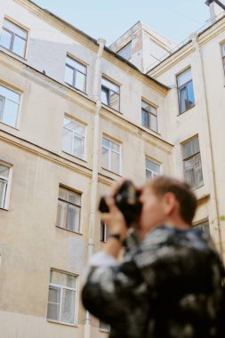 Adam, eski, uzun binanın dış yüzünü eski bir kamerayla fotoğraflıyor. Önplanda bulanık bir etki bırakıyor. Şehir sahnesine sanatsal bir dokunuş ekliyor.