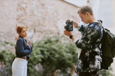 Genç yetişkin, arka planda başka bir bireyle modern kamerayı tutuyor duvara yaslanıyor deneğe odaklanıyor teknolojiyi kullanarak açık hava fotoğrafı yaratıyor.