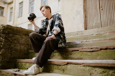 Genç, beyaz bir adam, elinde kamerayla dışarıda oturuyor, günlük giysiler giyiyor. Arka planda görünen tarihi yapı cephesi, kentsel bir çevre izlenimi veriyor