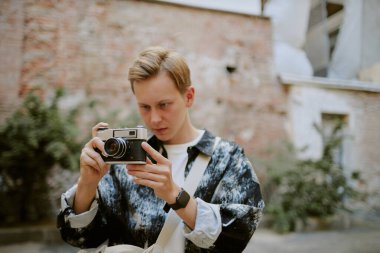 Kafkasyalı genç bir adam klasik kamerayı dışarı ayarlıyor, yeşil ve kırsal duvarların arkasında şehir ortamında çekim için hazırlanıyor.