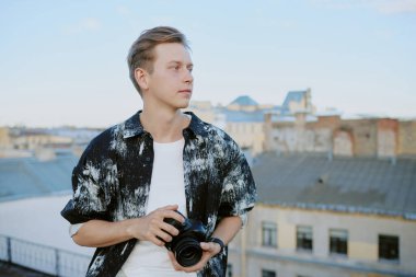 Beyaz adam elinde kamera ile şehir çatısı arka planında berrak gökyüzü altında gündüz saatlerinde binaların detaylarını yakalıyor.