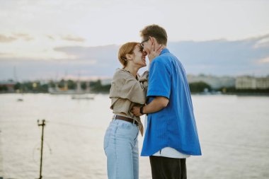 Günbatımında deniz kenarında samimi bir anı paylaşan genç bir çift, arka planda şehir silüeti. Gündelik kıyafetler ve doğal ortam romantik bir hava katıyor.
